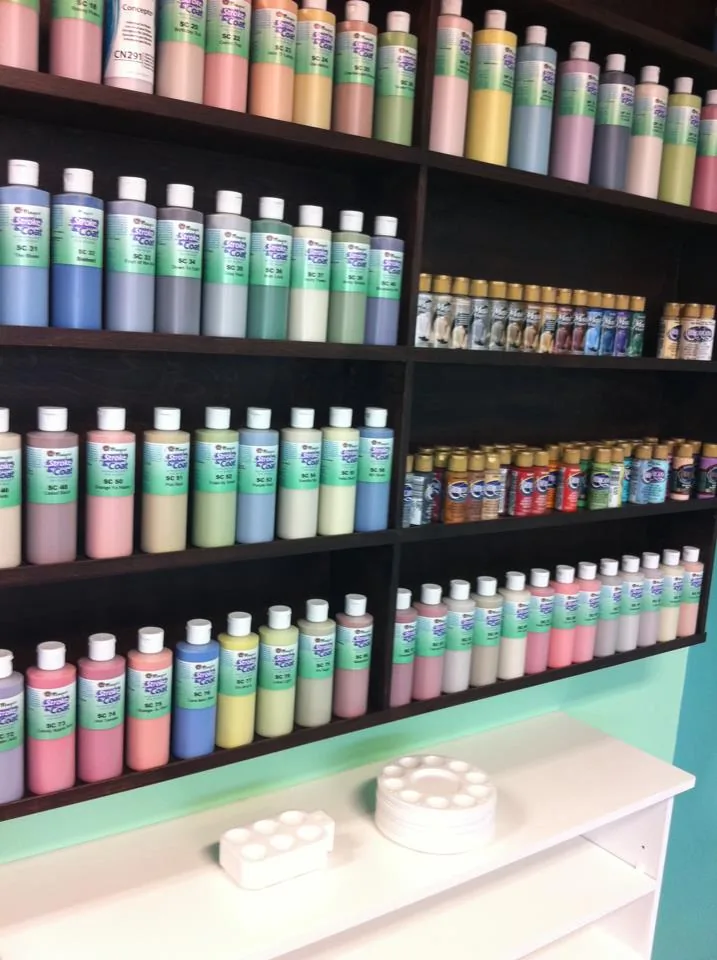Shelves of ceramic paint bottles in many colours, with paint palettes on a counter below