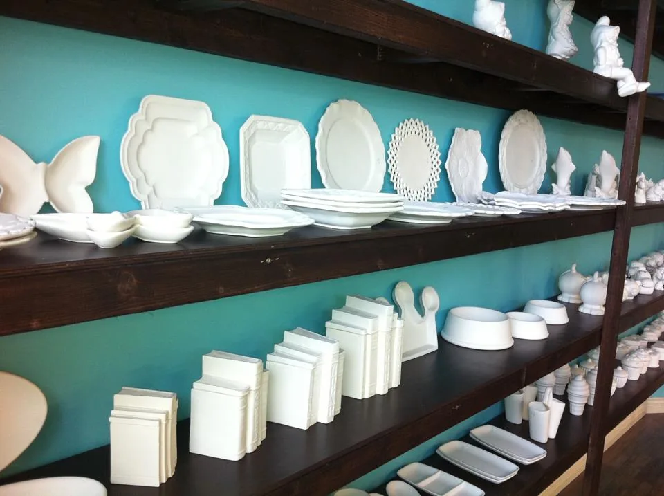 Unpainted white ceramic plates, bowls and figures on dark wooden shelves against a turquoise wall