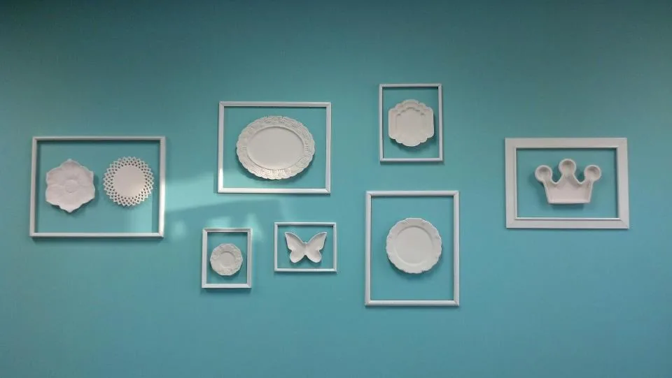 White ceramic plates, a butterfly and a crown displayed inside white frames on a turquoise wall